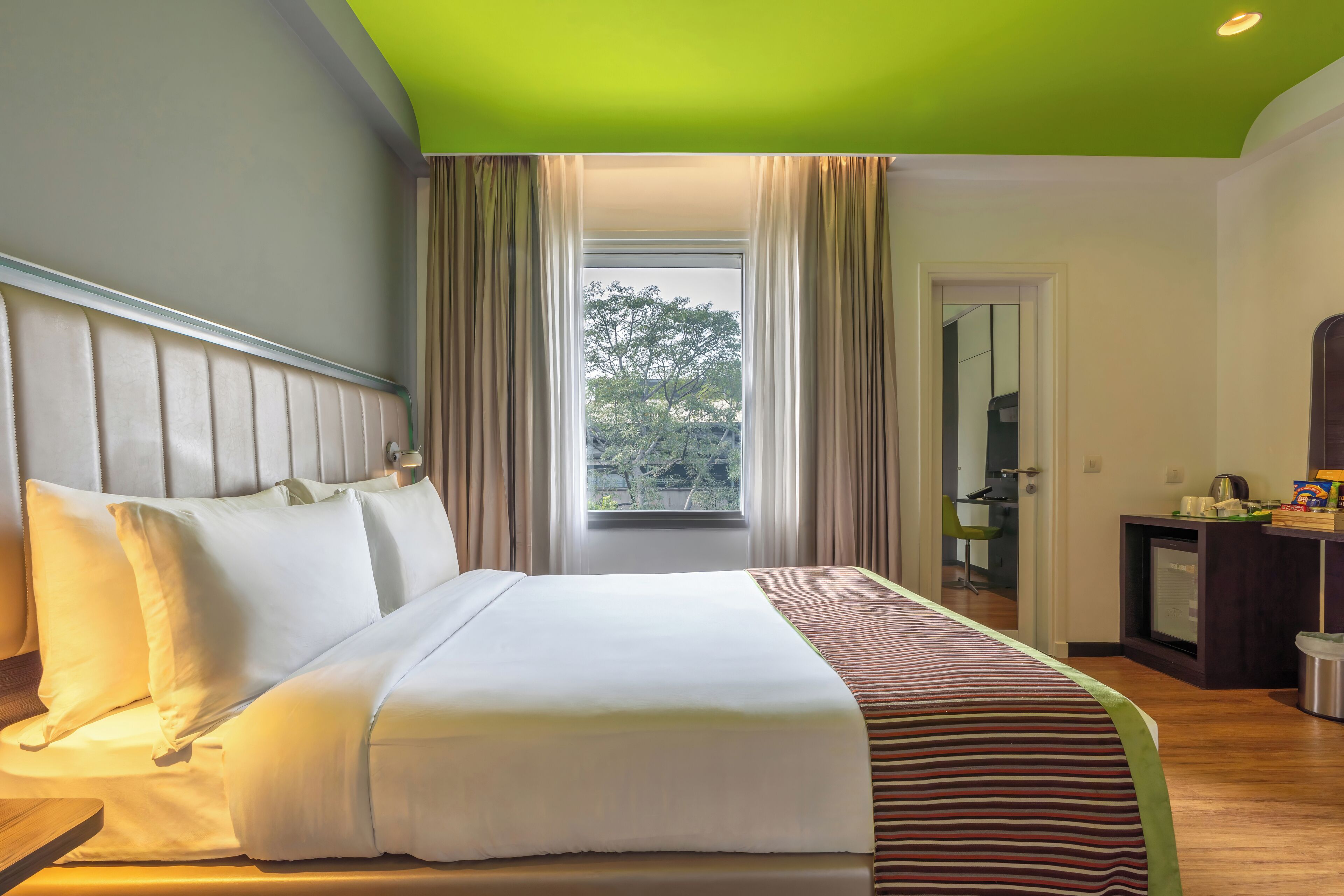 Habitación de hotel con una cama grande, una ventana con cortinas, un escritorio y una silla, y vista a árboles desde afuera.