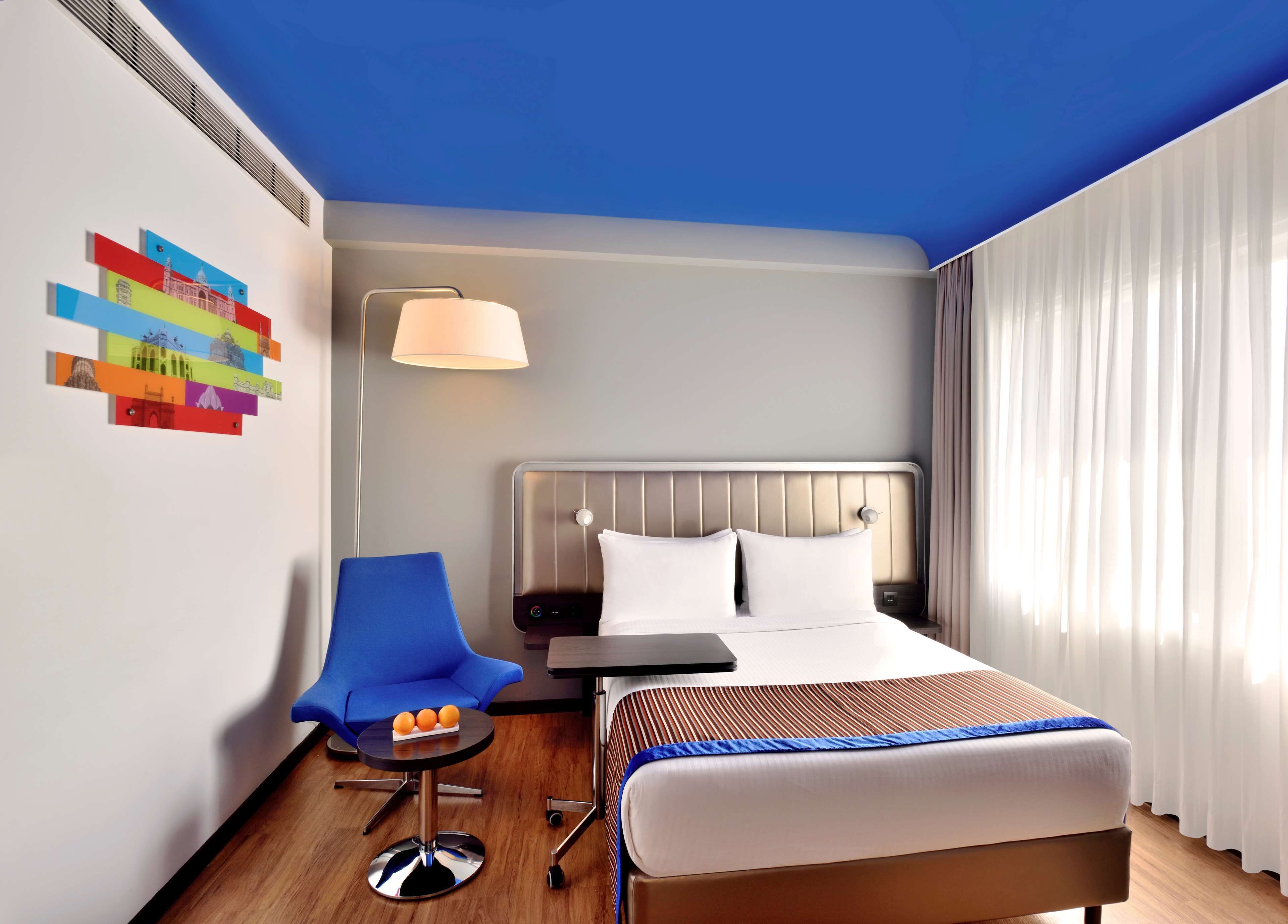 A hotel room with a bed, a chair, a small table with oranges, a lamp, and a wall with colorful artwork.