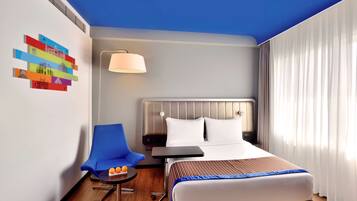 A hotel room with a bed, a chair, a small table with oranges, a lamp, and a wall with colorful artwork.