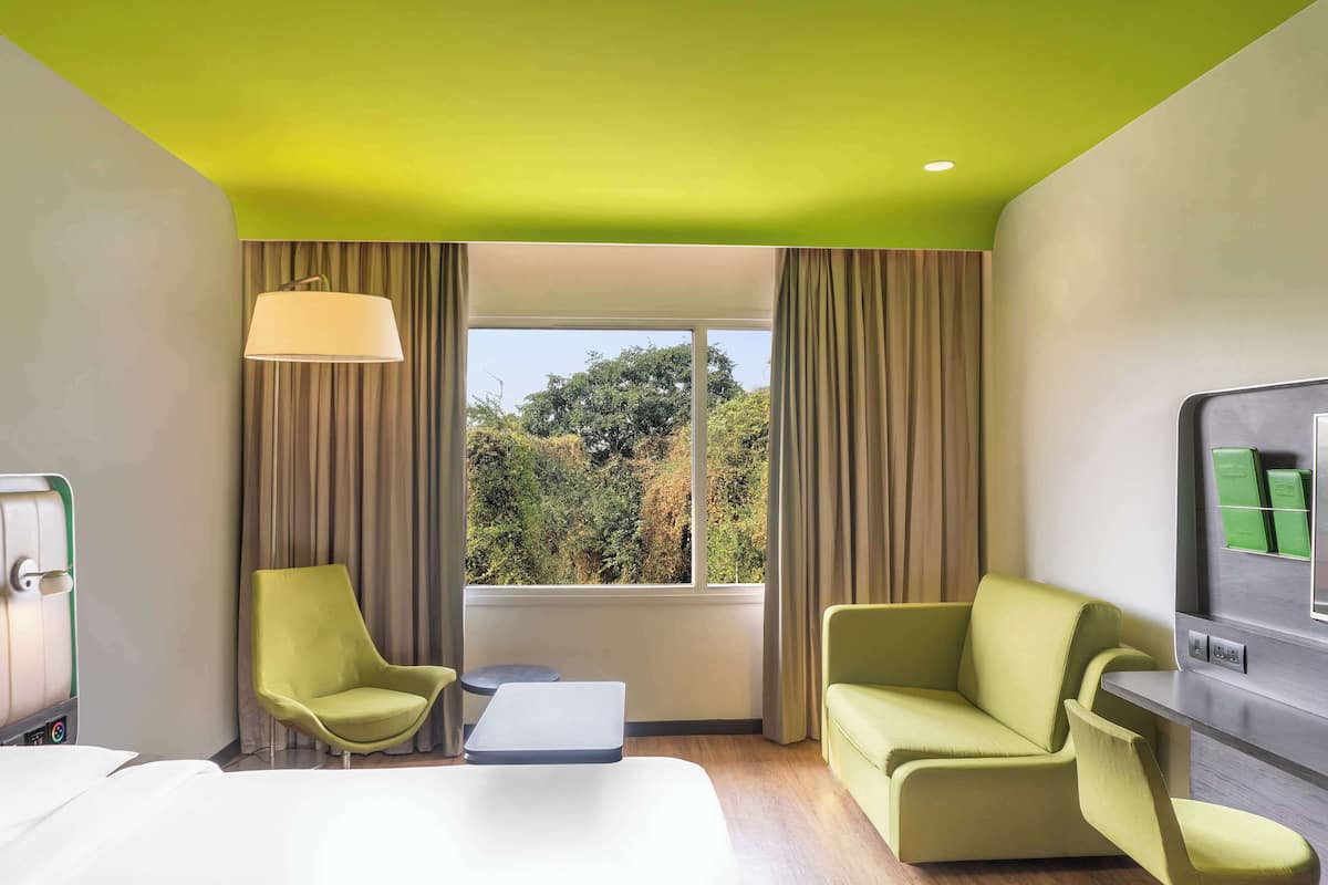 A hotel room with a bed, two green armchairs, a small table, a lamp, a TV, and a window with curtains.