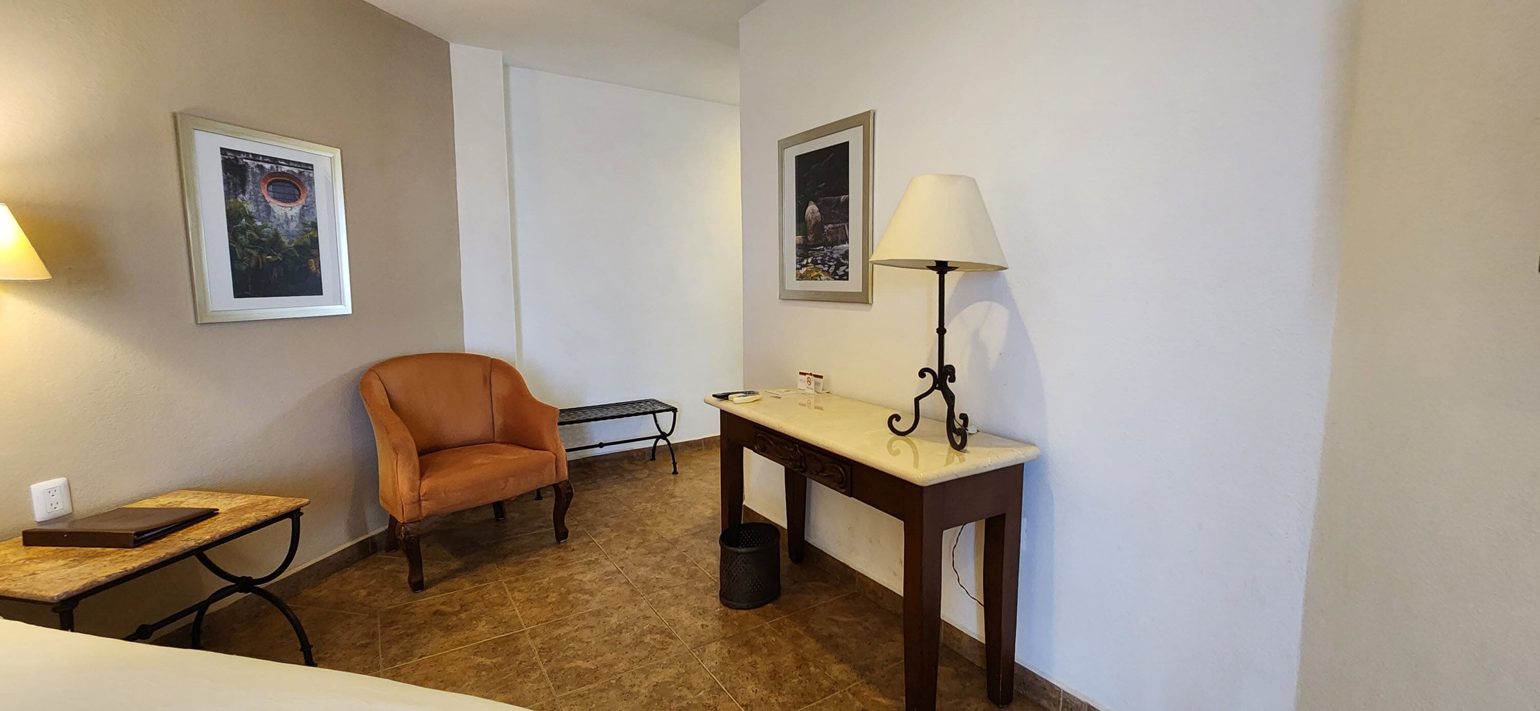 Una habitación de hotel con una sola silla naranja, una mesita con una lámpara, un cuadro en la pared y un cesto de basura.