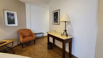 Una habitación de hotel con una sola silla naranja, una mesita con una lámpara, un cuadro en la pared y un cesto de basura.