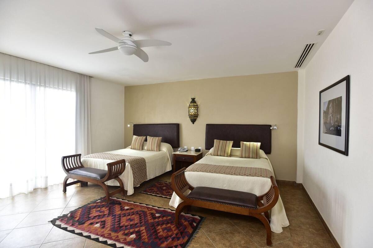 Ein Hotelzimmer mit zwei Betten, einem Deckenventilator, einem Nachttisch, einem Sessel, einem Teppich und einer Wandlampe.