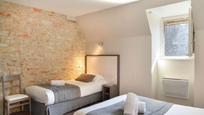A hotel room with a stone wall, two beds, a chair, a nightstand, and a window with an air conditioning unit. - Le Puits Doré – Hôtel Restaurant Logis (Richelieu)