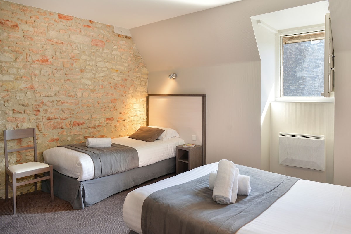 Habitación de hotel con pared de piedra, dos camas, una silla, una mesita de noche y una ventana con aire acondicionado.