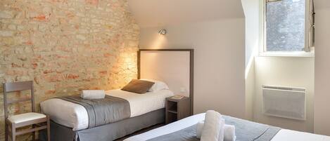 A hotel room with a stone wall, two beds, a chair, a nightstand, and a window with an air conditioning unit.