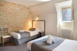 Une chambre d’hôtel avec un mur en pierre, deux lits, une chaise, une table de chevet et une fenêtre équipée d’un appareil de climatisation.