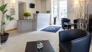 A reception area with a reception desk, a potted plant, a seating area with a black leather chair, and a blue mat with a wheelchair symbol. - Le Puits Doré – Hôtel Restaurant Logis (Richelieu)