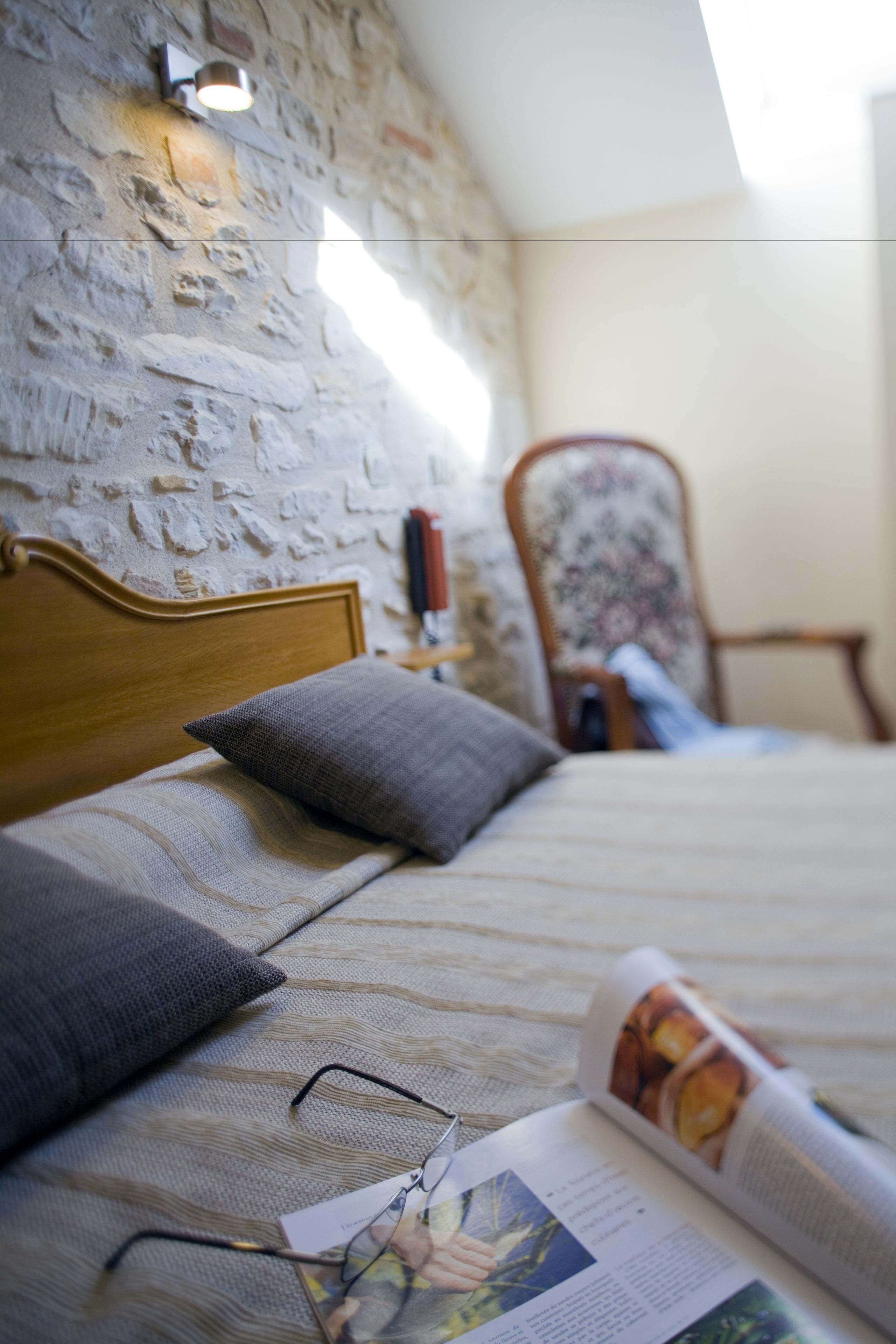 A bedroom with a stone wall, a wooden headboard, a bed with grey bedding, a pair of glasses, and a magazine on the bed.