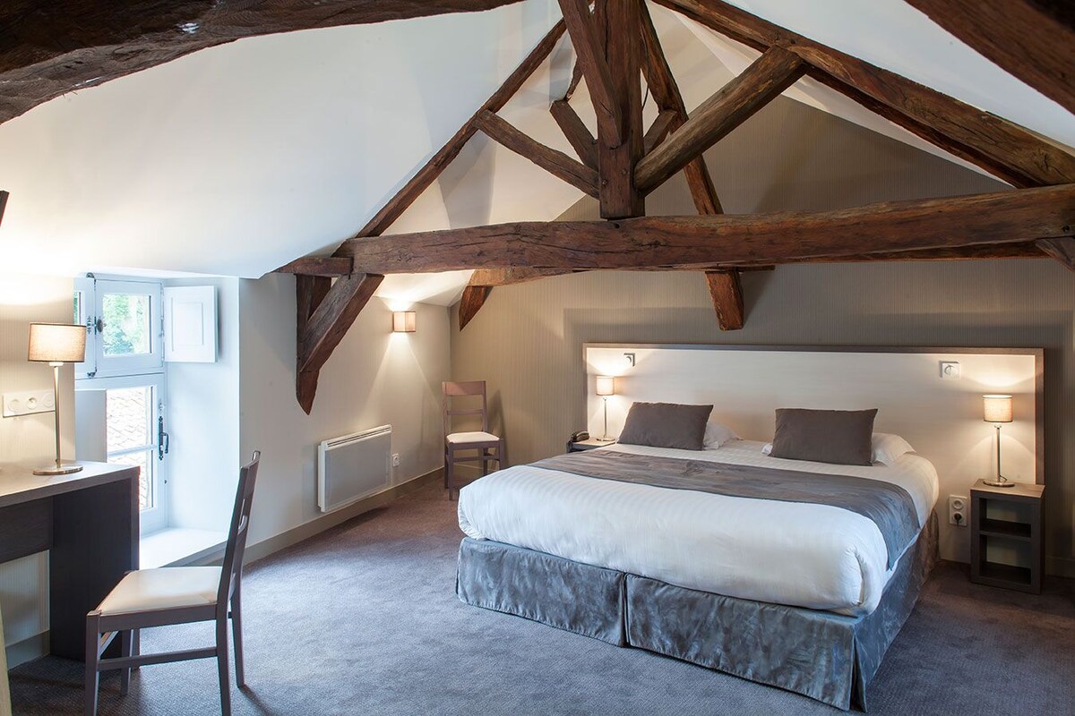 A bedroom with a large bed, a desk, and a chair. There are two bedside lamps and a window with a view.