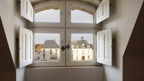 A room with a large arched window overlooking a building with multiple stories and a bell tower. - Le Puits Doré – Hôtel Restaurant Logis (Richelieu)