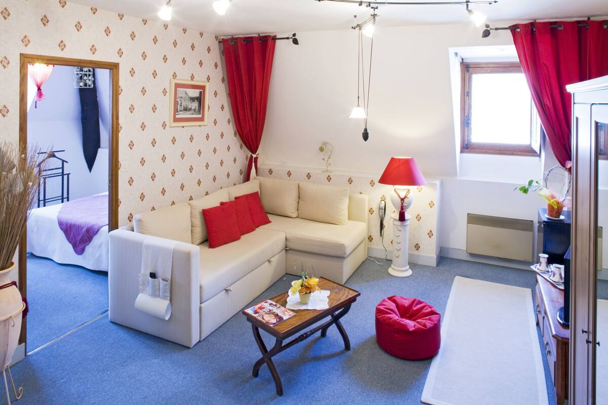 Un salón con un sofá color beige, cojines rojos, una mesa de centro de madera y un sillón de peluche rojo.