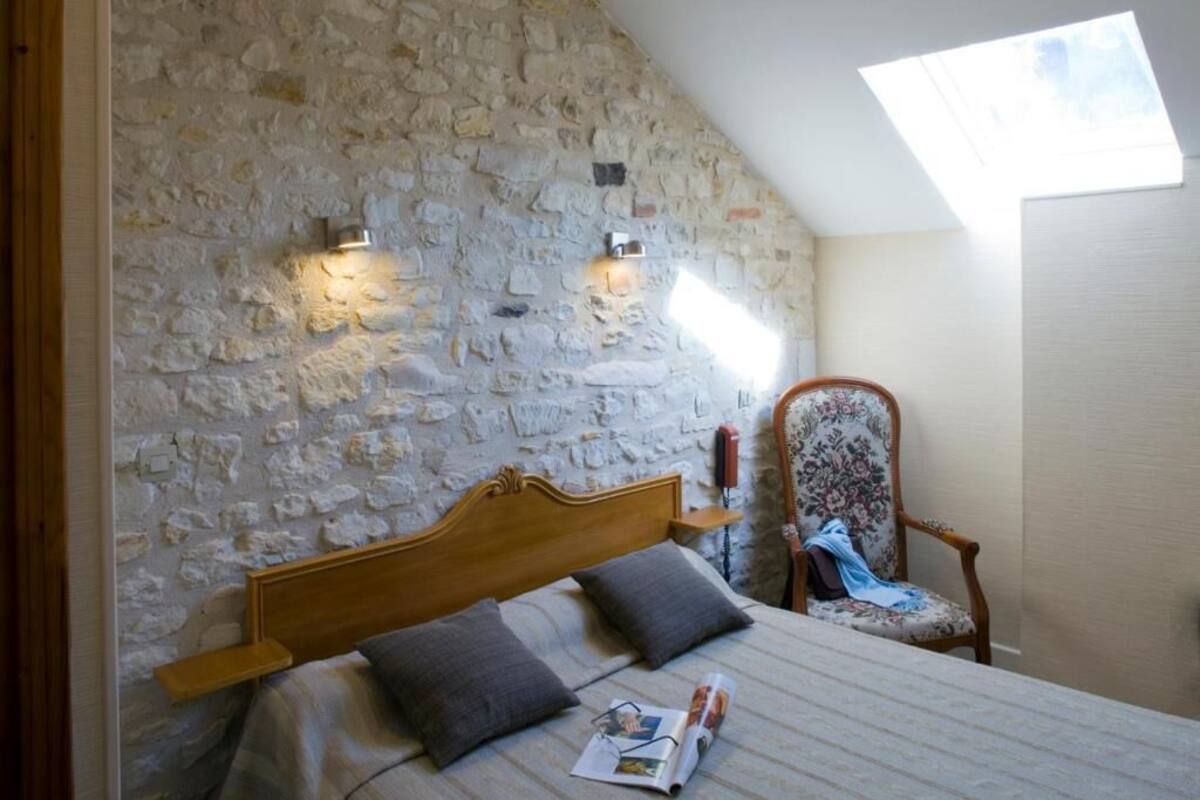 Un dormitorio con pared de piedra, cama de madera, una silla y tragaluz.