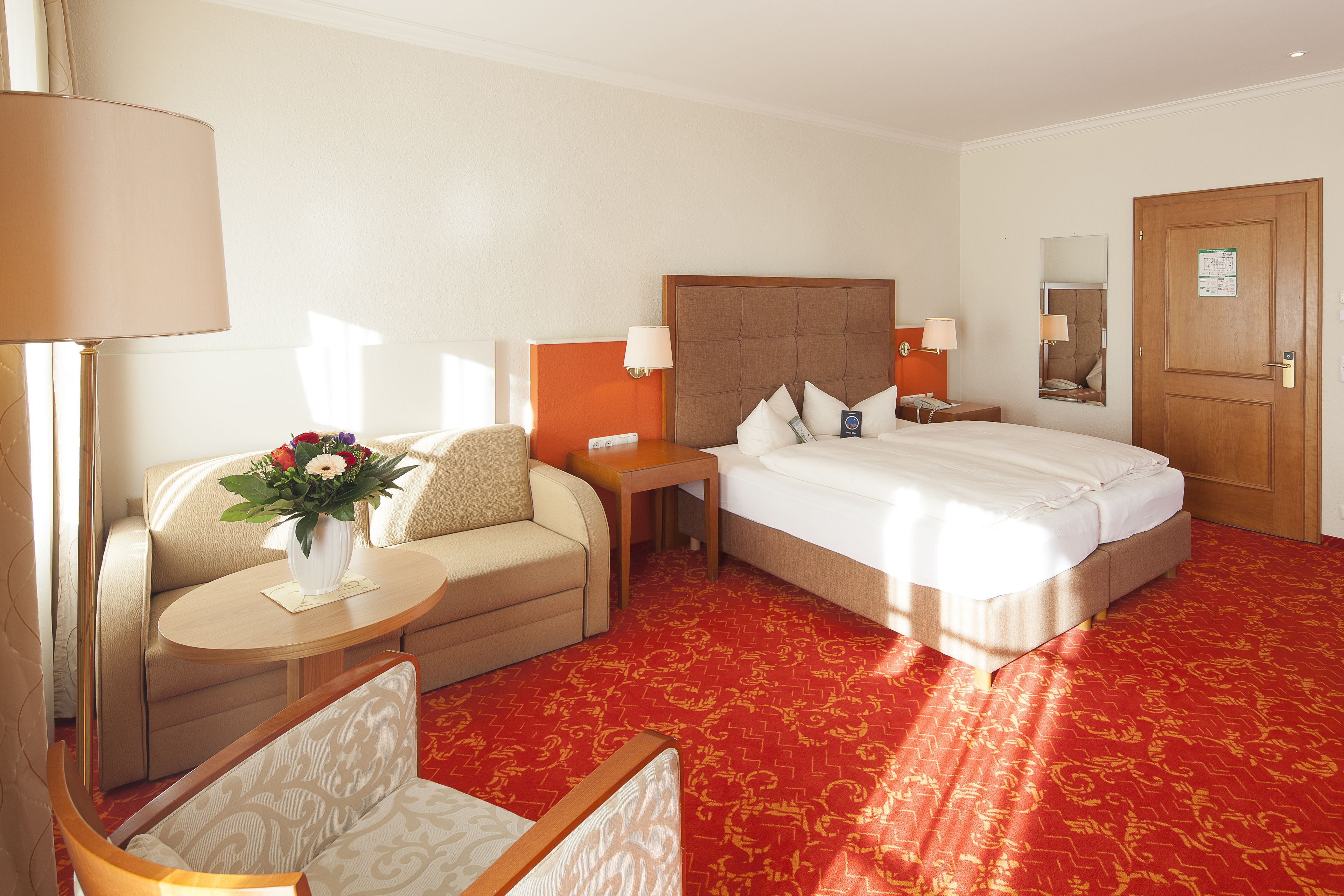 A hotel room with a bed, a sofa, a small table with a vase of flowers, and a nightstand with a lamp.