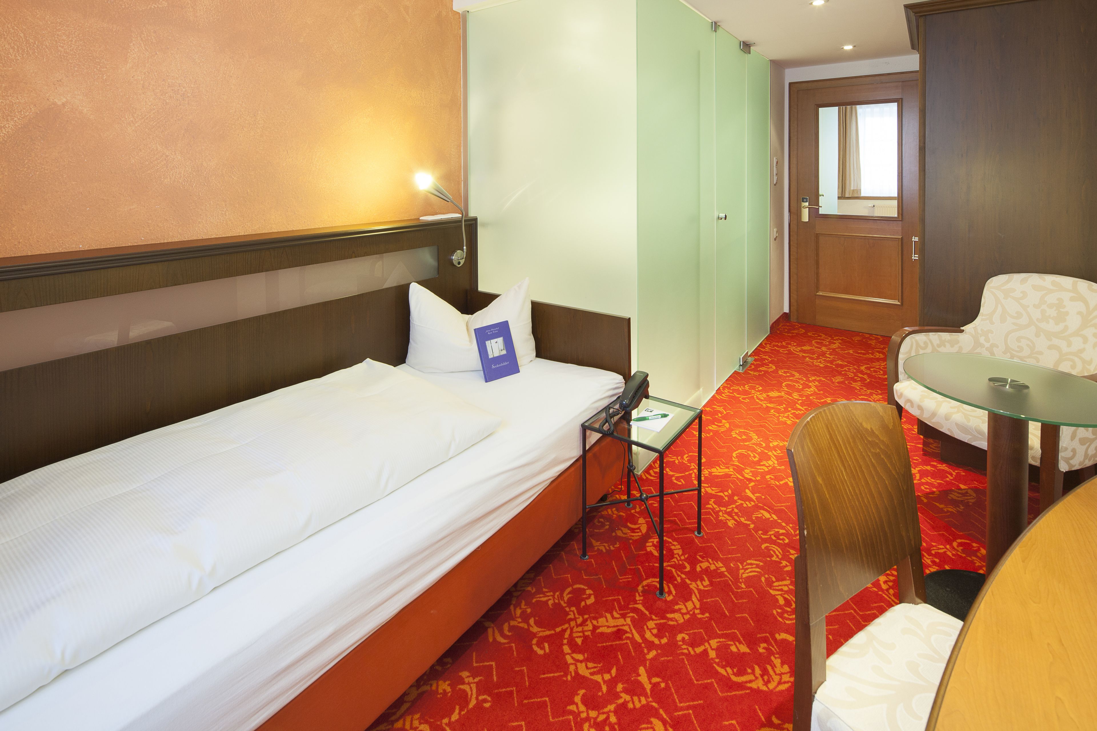 Una habitación de hotel con una cama individual, una mesita, una silla y un panel de vidrio.