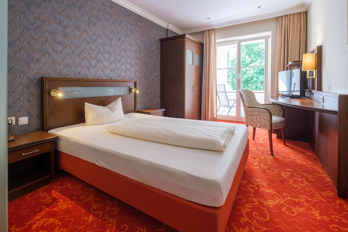 A hotel room with a large bed, a desk with a lamp, a wardrobe, and a chair.