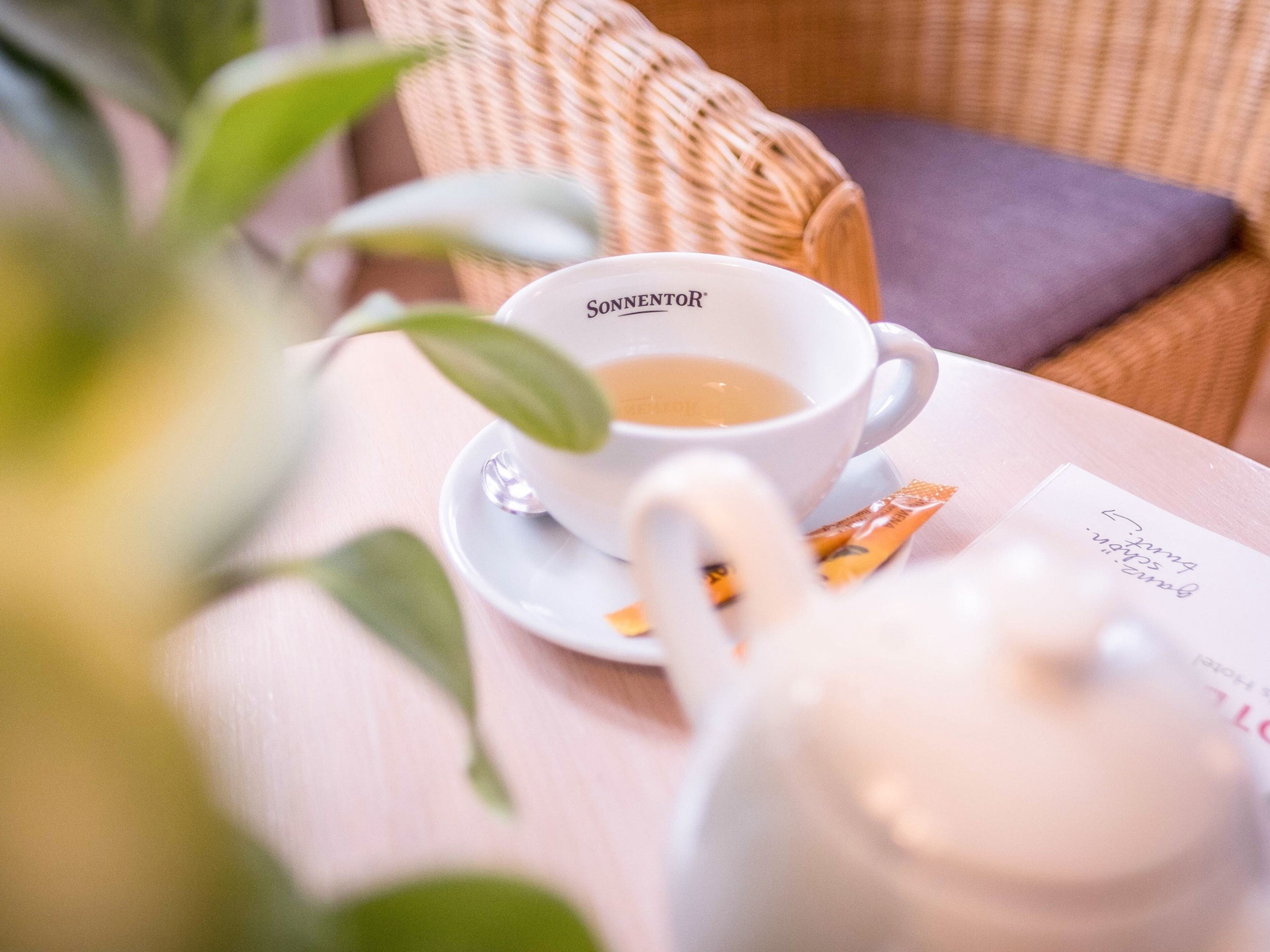 Una tetera blanca y una taza blanca con la marca 'SONNENTOR', sobre una mesa con un cojín y una bolsita de té.