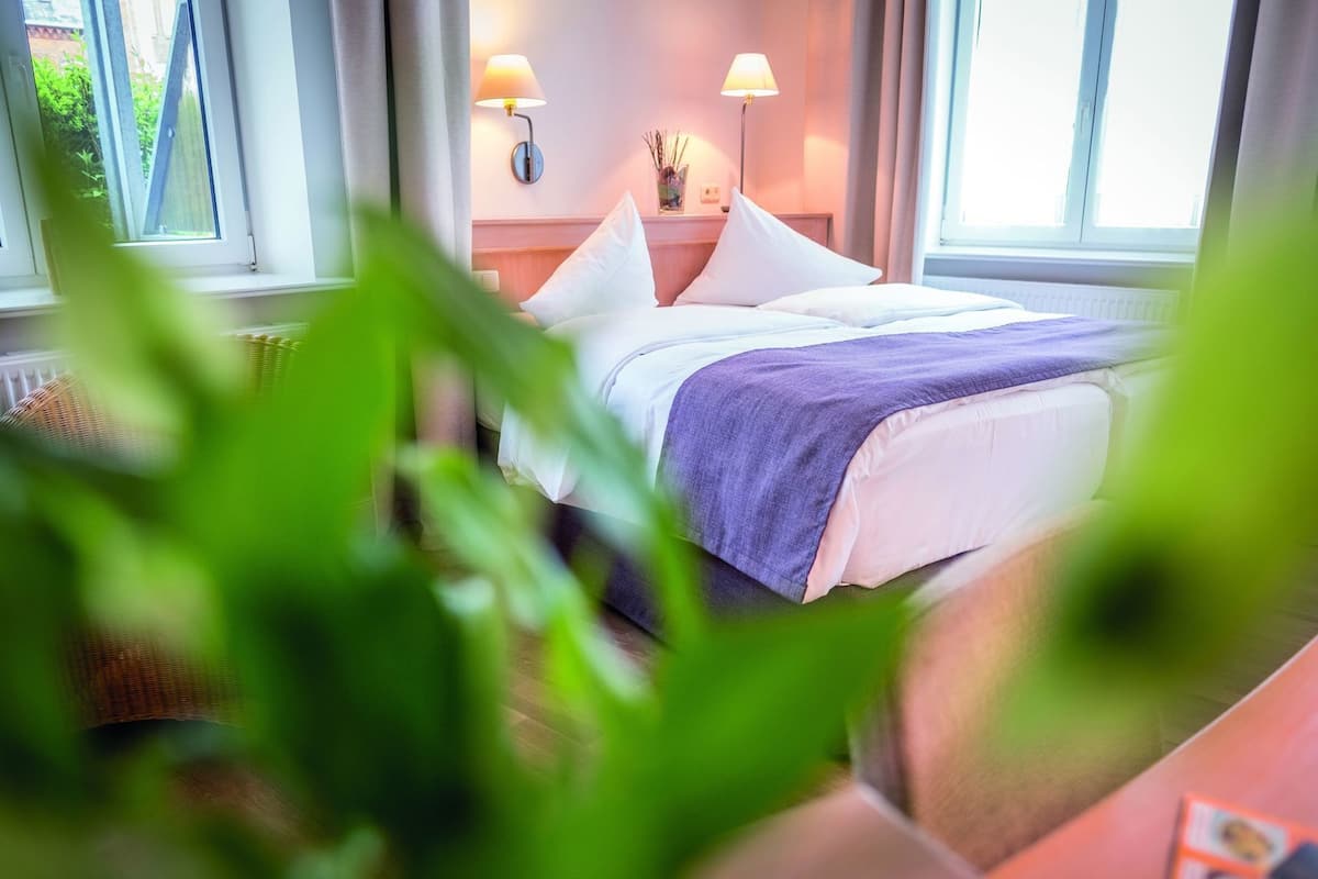 A hotel room with a bed, two bedside lamps, a window, and a view of greenery outside.