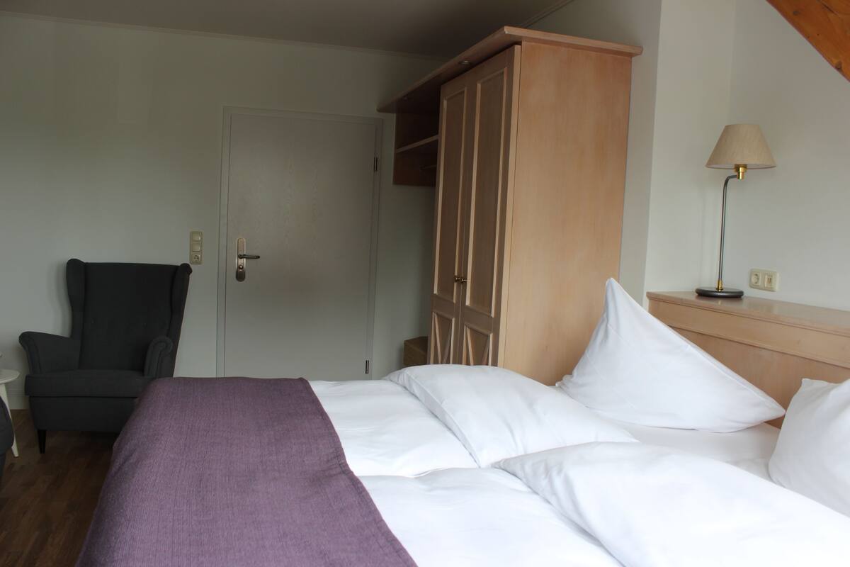 A hotel room with a bed, a chair, a wardrobe, and a nightstand with a lamp.