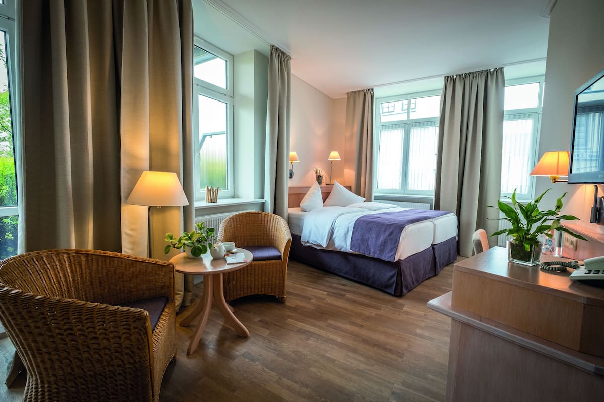 A hotel room with a bed, two chairs, a small table with a lamp, and a desk with a phone.