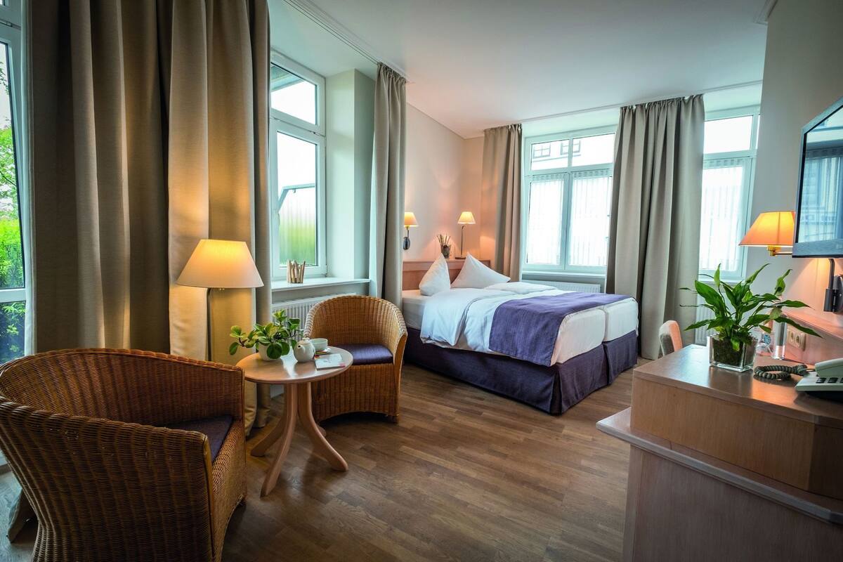 A hotel room with a bed, a desk, a chair, a small table with a plant, and a window with curtains.