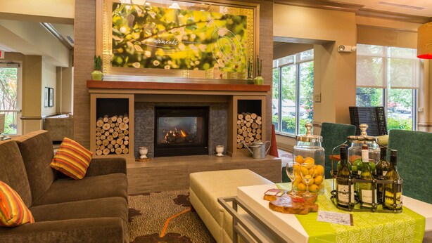 A cozy living room with a fireplace, a green table with bottles, and a couch with colorful pillows. - Hilton Garden Inn Charlottesville (Charlottesville)