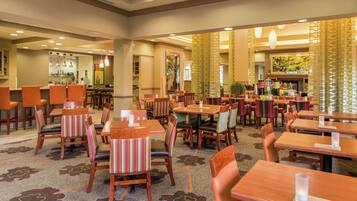 A hotel dining area with multiple tables and chairs, a kitchen in the background, and decorative elements like curtains and plants.