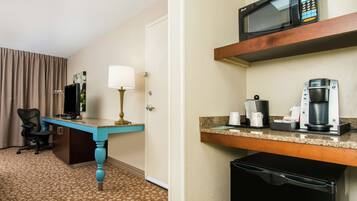 A hotel room with a desk, a chair, a lamp, a television, a microwave, a coffee maker, and a trash bin.