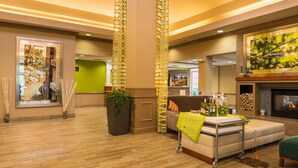 A lobby with a wooden floor, a sofa, a cart with bottles, a fireplace, and a green wall. - Hilton Garden Inn Charlottesville (Charlottesville)