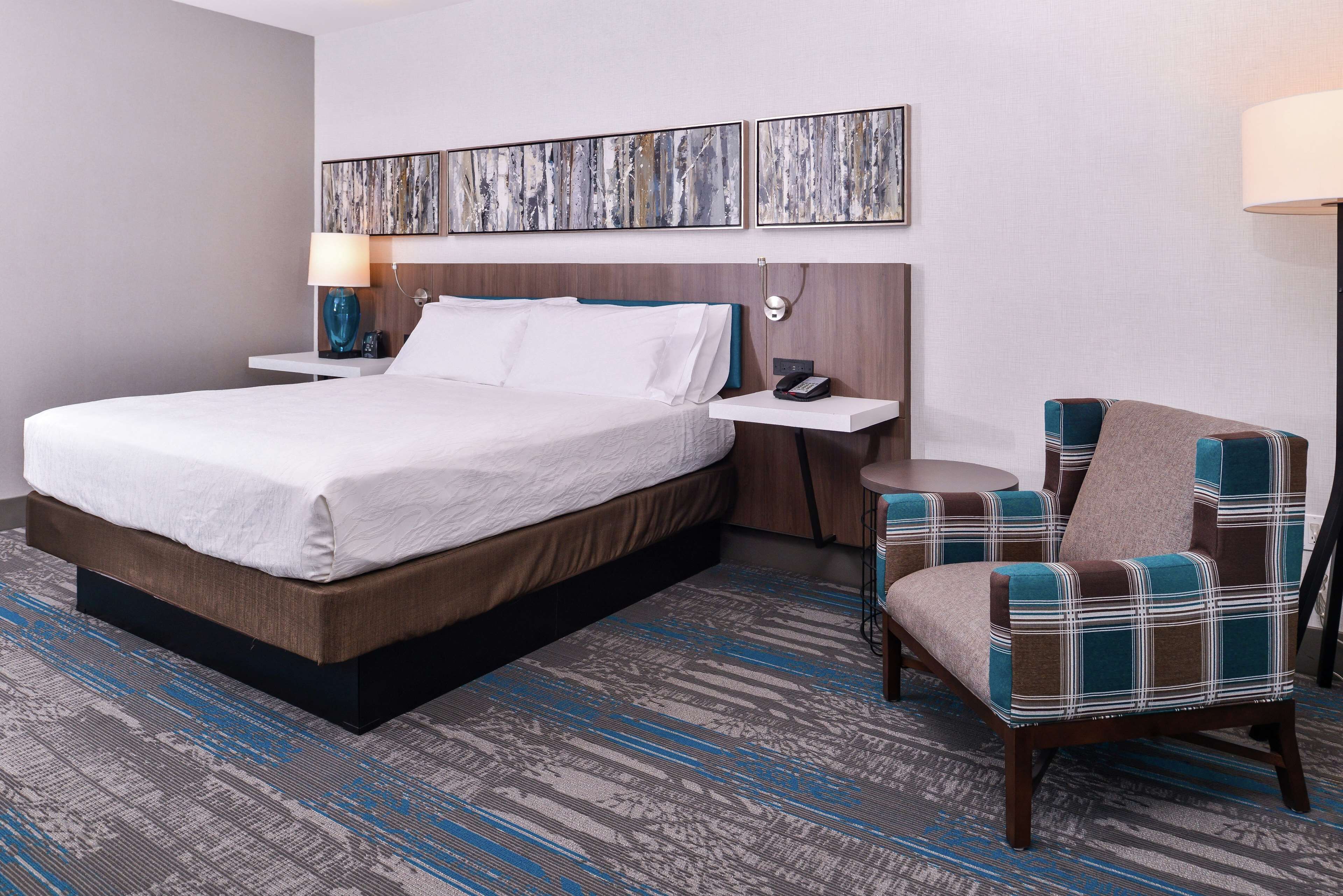 A hotel room with a large bed, a bedside table with a lamp, a chair, and two abstract paintings on the wall.