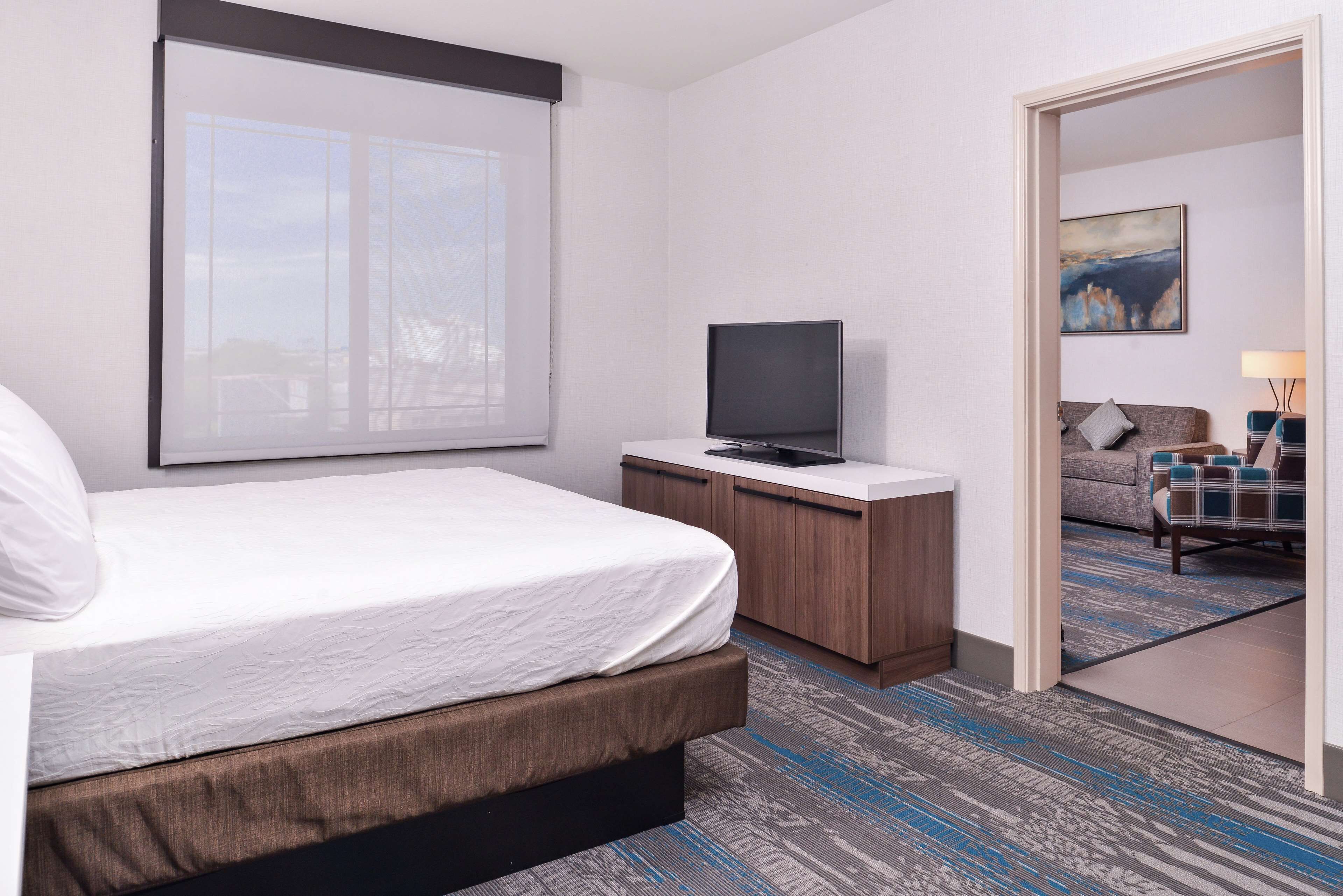 A hotel room with a bed, a TV on a wooden cabinet, a painting on the wall, and a view through an open door.