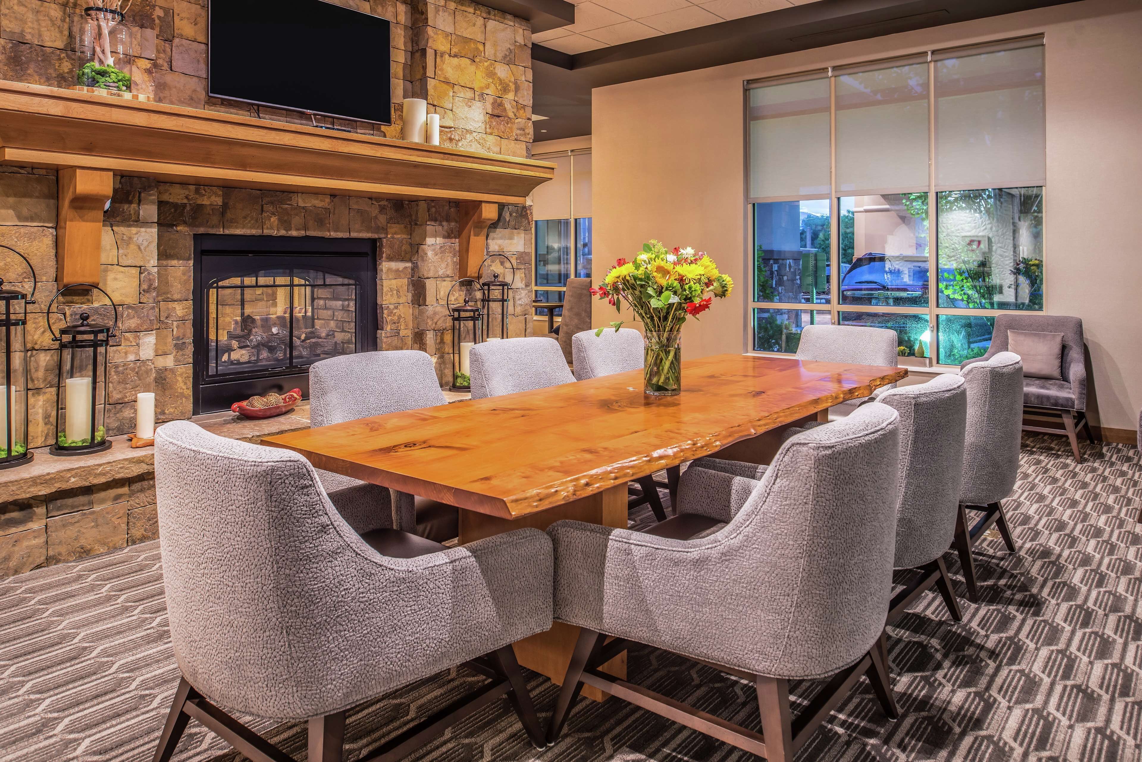 A conference room with a long wooden table, six upholstered chairs, a stone fireplace, and a large window offering a view of the outdoors.