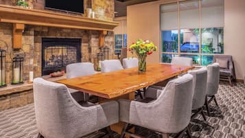 A conference room with a long wooden table, six upholstered chairs, a stone fireplace, and a large window offering a view of the outdoors.