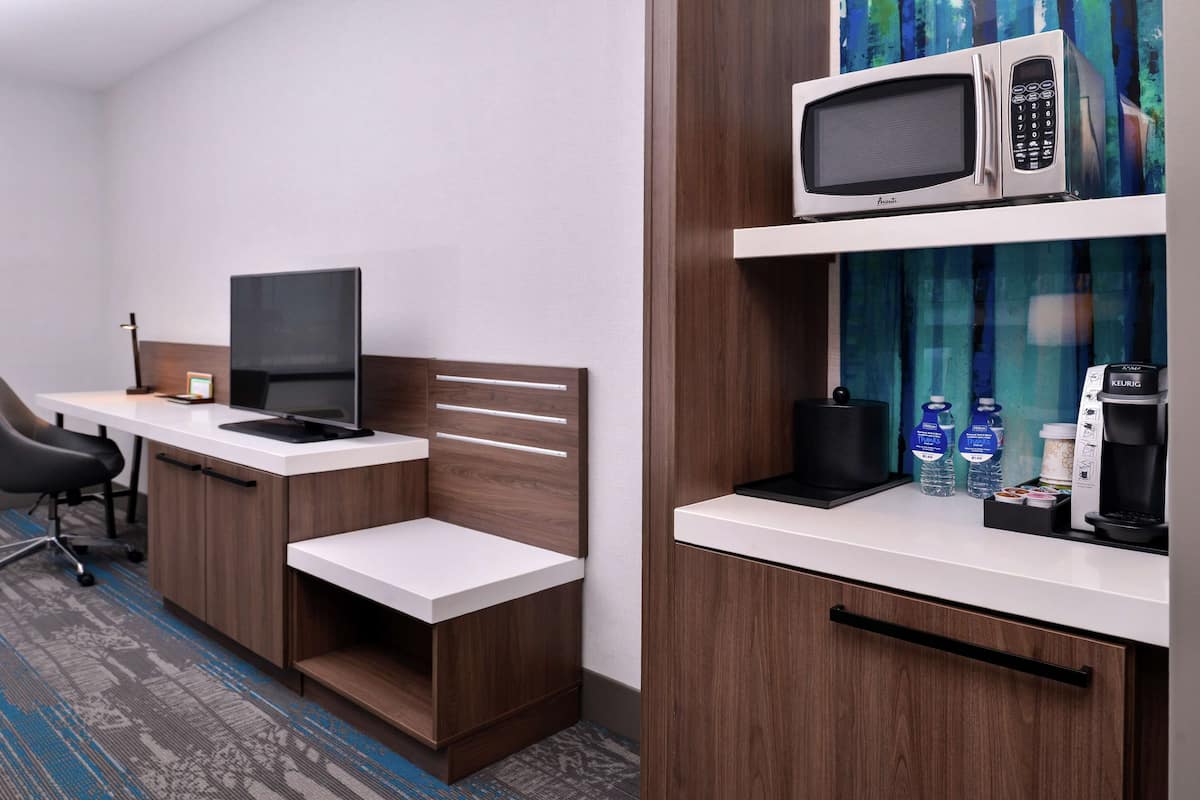 Ein Hotelzimmer mit Schreibtisch, Computer, Stuhl, Bett, Mikrowelle, Kaffeemaschine und Wasserflaschen.
