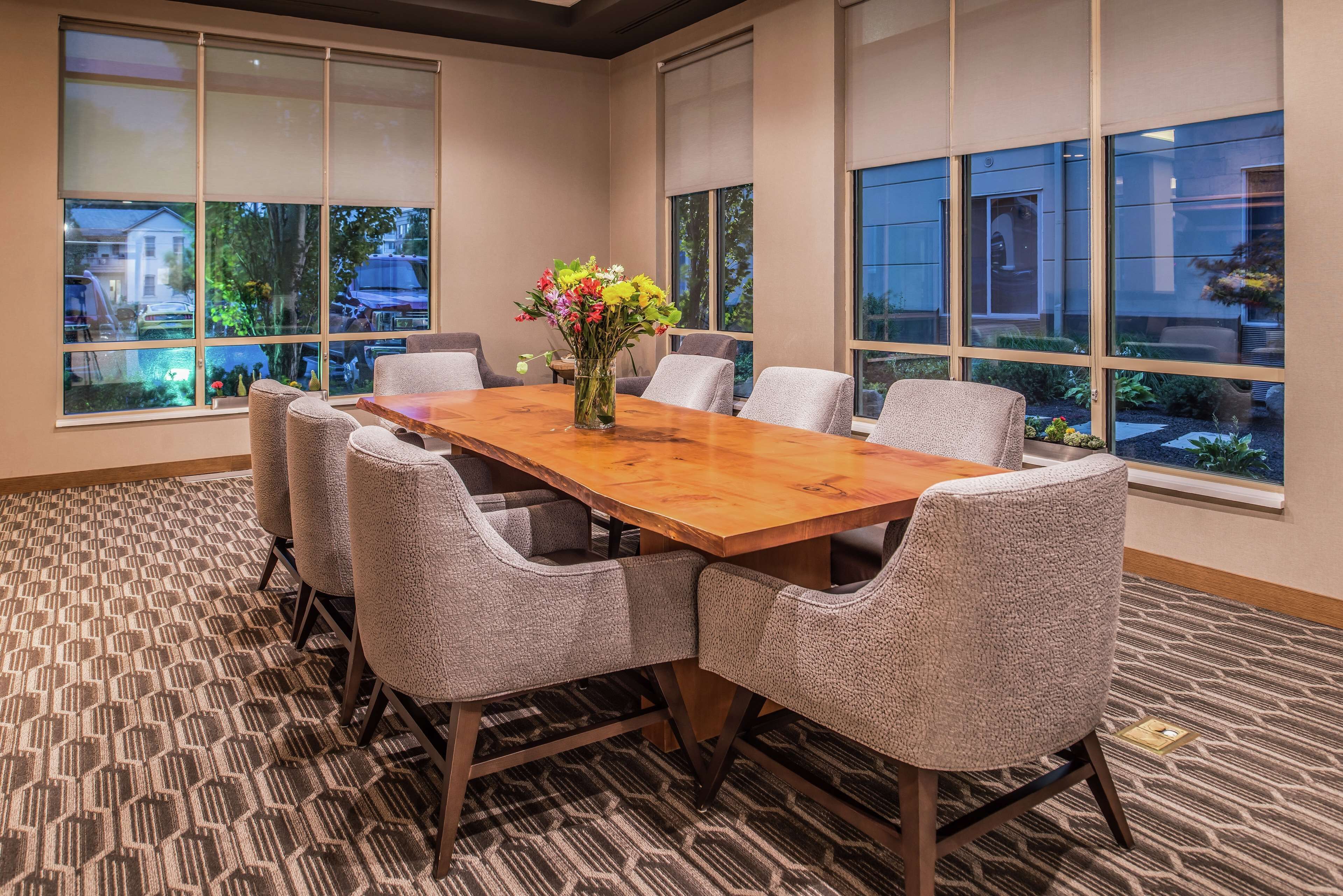 A conference room with a wooden table, six upholstered chairs, and large windows offering a view of the outdoors.