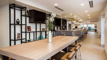 A modern dining area with a long wooden table, black shelving units, and a television mounted on the wall.