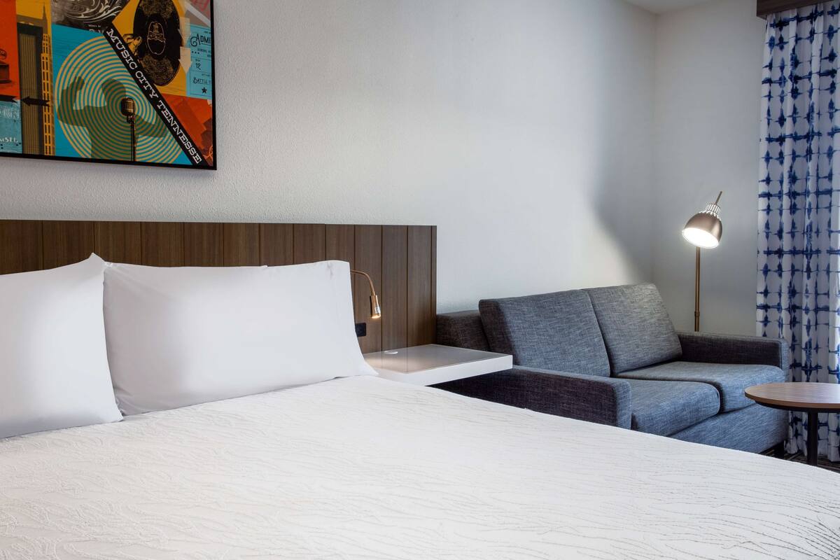 A hotel room with a bed, a gray sofa, a small table, a lamp, and a wall-mounted painting.
