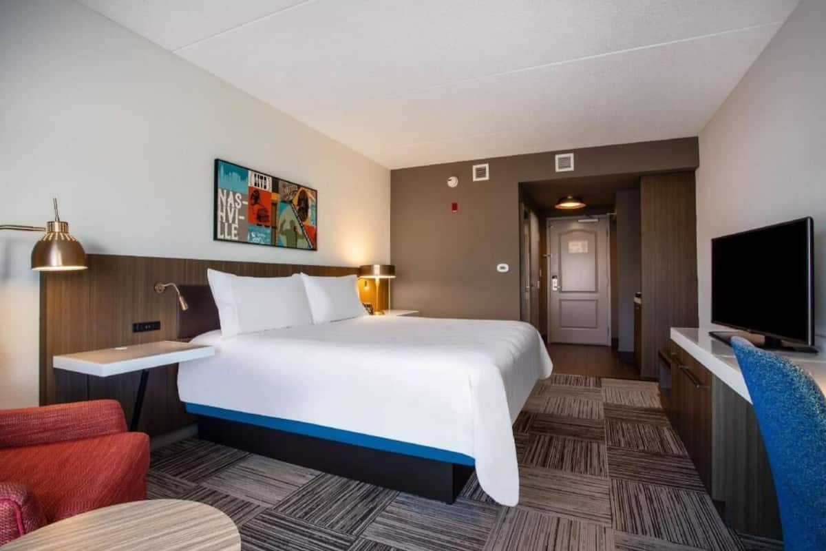 A hotel room with a bed, bedside tables, a TV, a painting, and a door leading to a bathroom.
