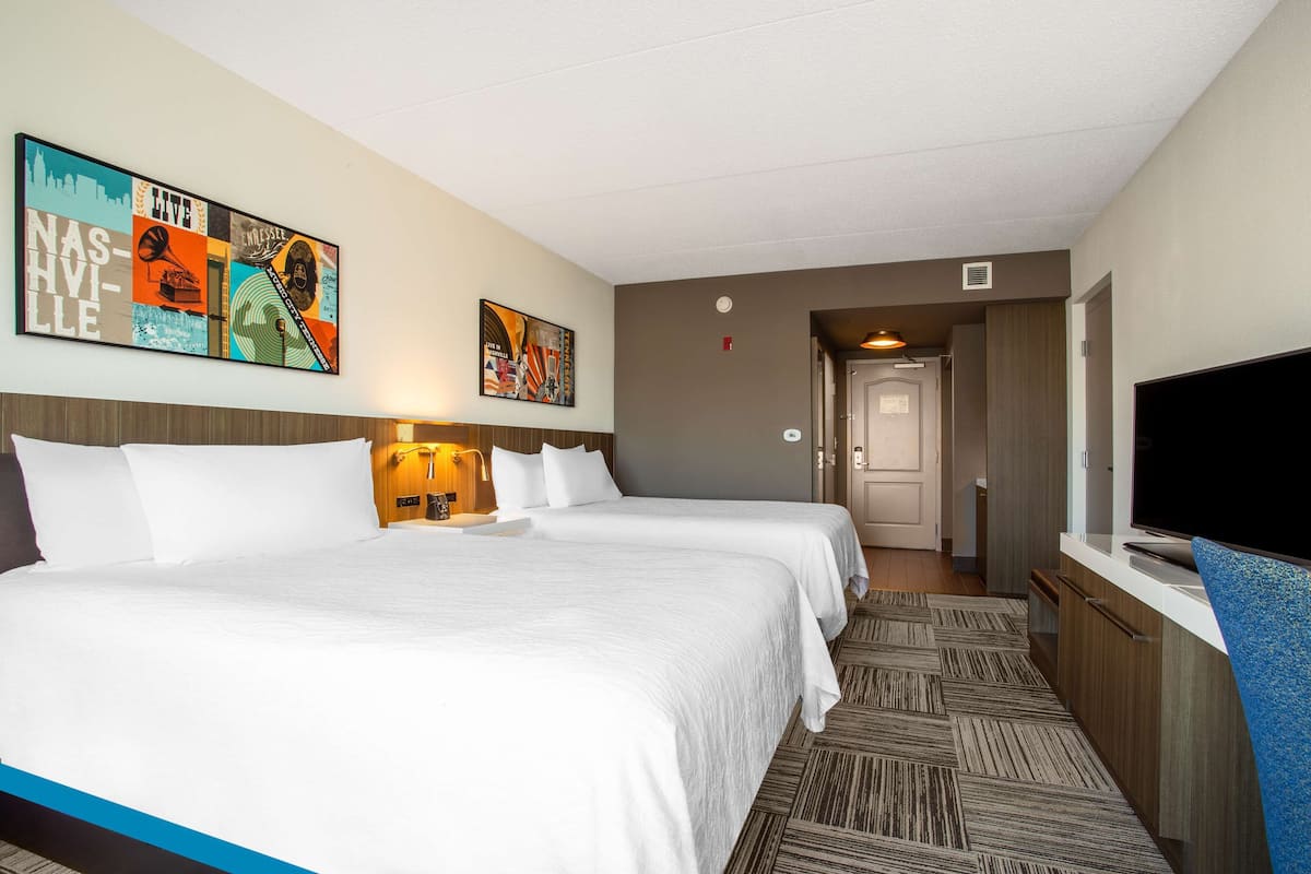 A hotel room with two beds, a TV, a painting on the wall, and a carpeted floor.