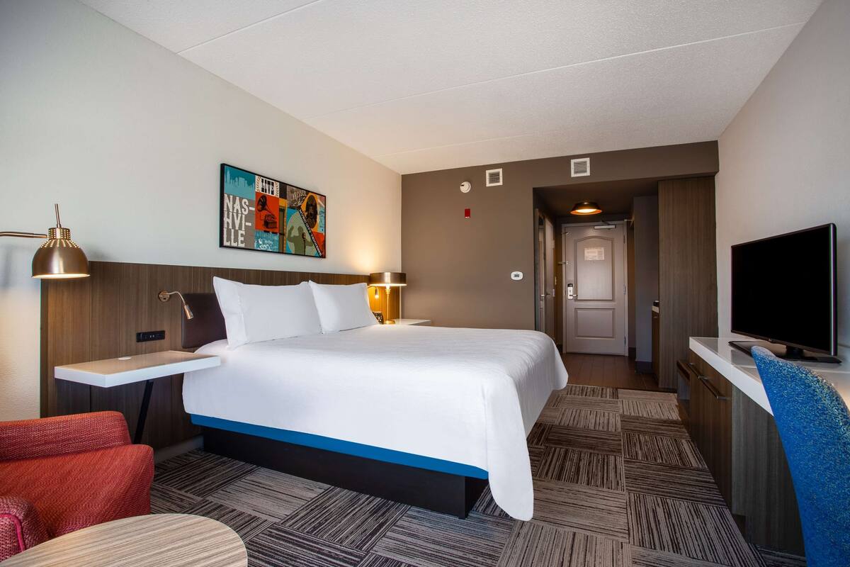 A hotel room with a bed, bedside tables, a TV, a painting, and a door leading to a bathroom.