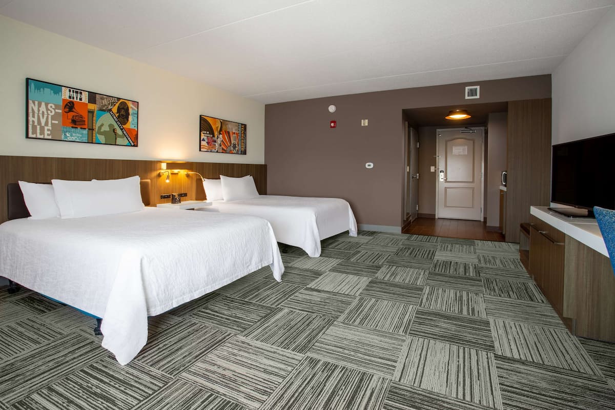 A hotel room with two beds, a desk, and a television. There are wall art pieces and a door leading to another room.