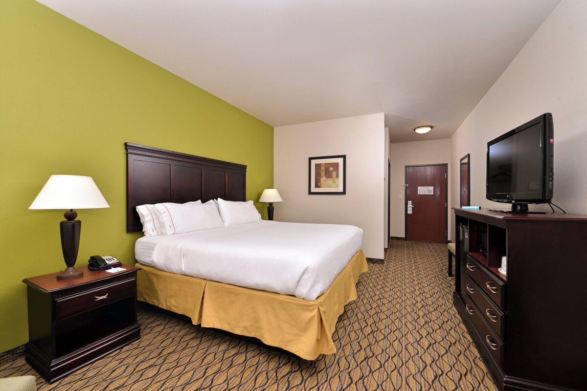 A hotel room with a bed, bedside lamps, a TV, a dresser, and a door leading to another room.