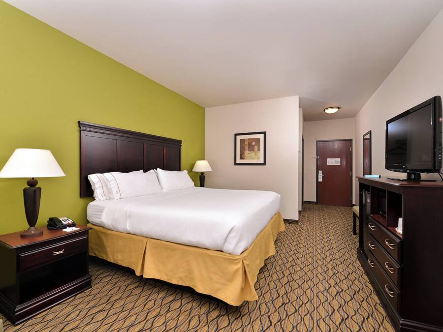 A hotel room with a bed, bedside lamps, a TV, a dresser, and a door leading to another room.