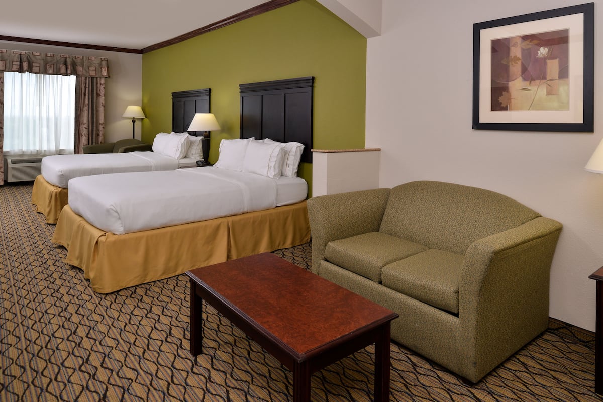 A hotel room with two beds, a green armchair, a wooden table, and a framed picture on the wall.