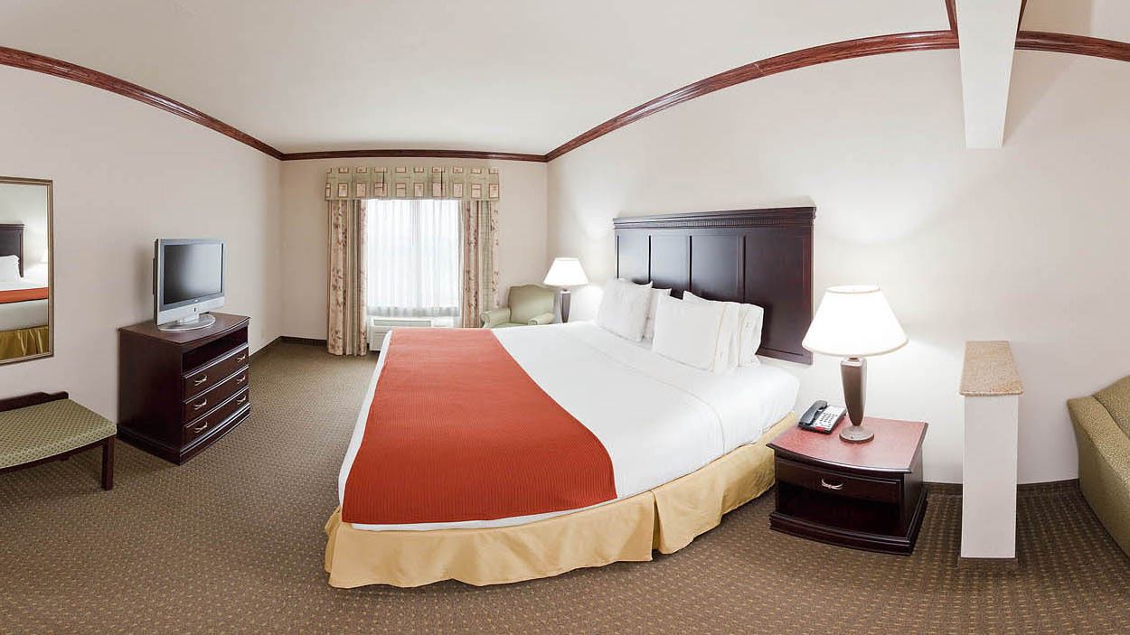 A hotel room with a large bed, a television on a dresser, a desk with a lamp, and a chair.