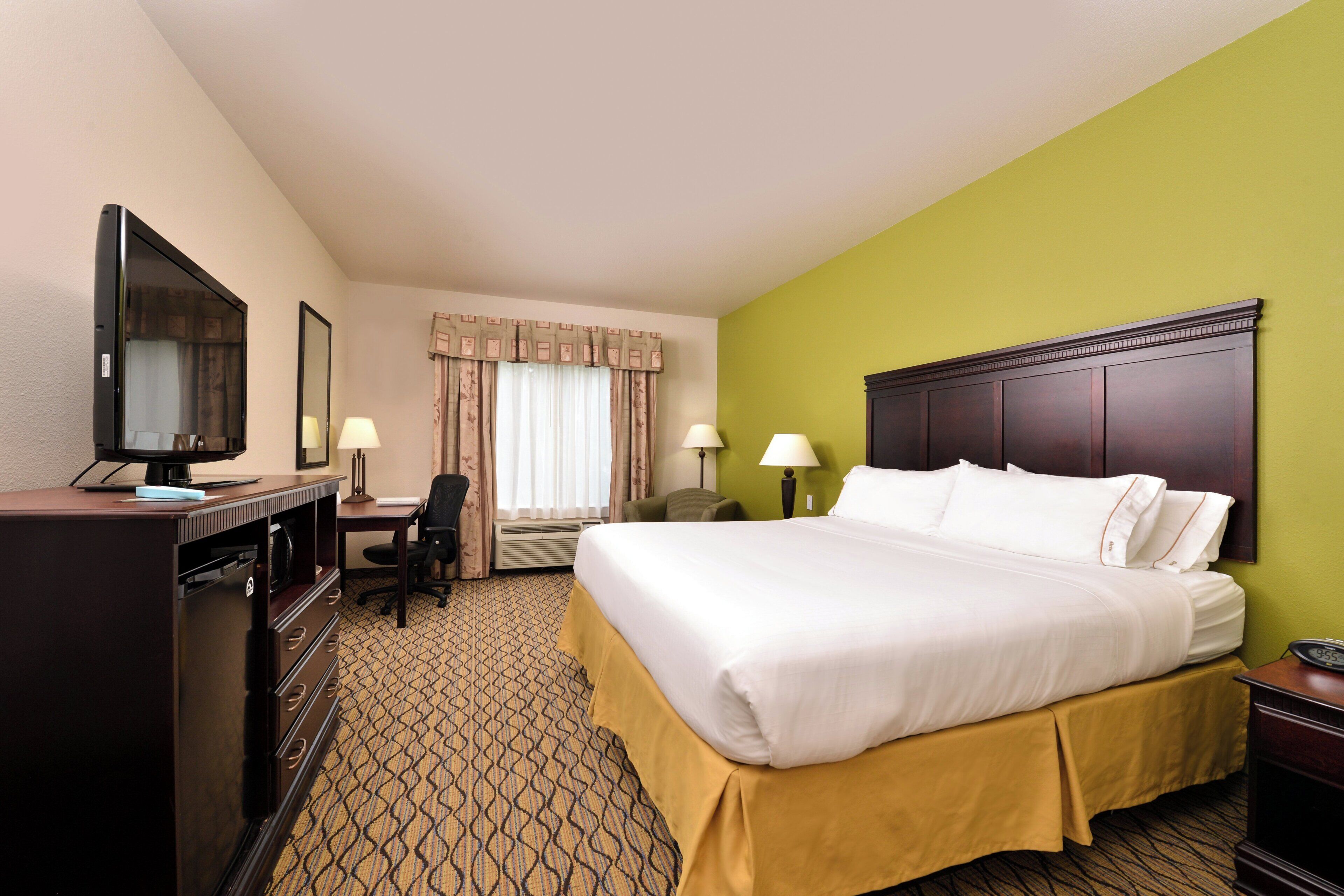 A hotel room with a bed, desk, TV, and a window with curtains.
