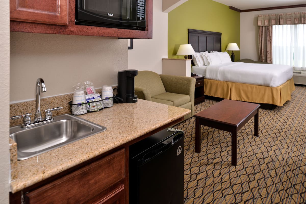 A hotel room with a kitchenette, including a sink, stove, and microwave. There is a bed with a nightstand, two lamps, and a window with curtains.
