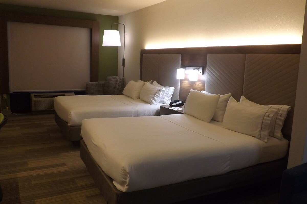 A hotel room with two beds, a desk, a lamp, and a view of the outside.