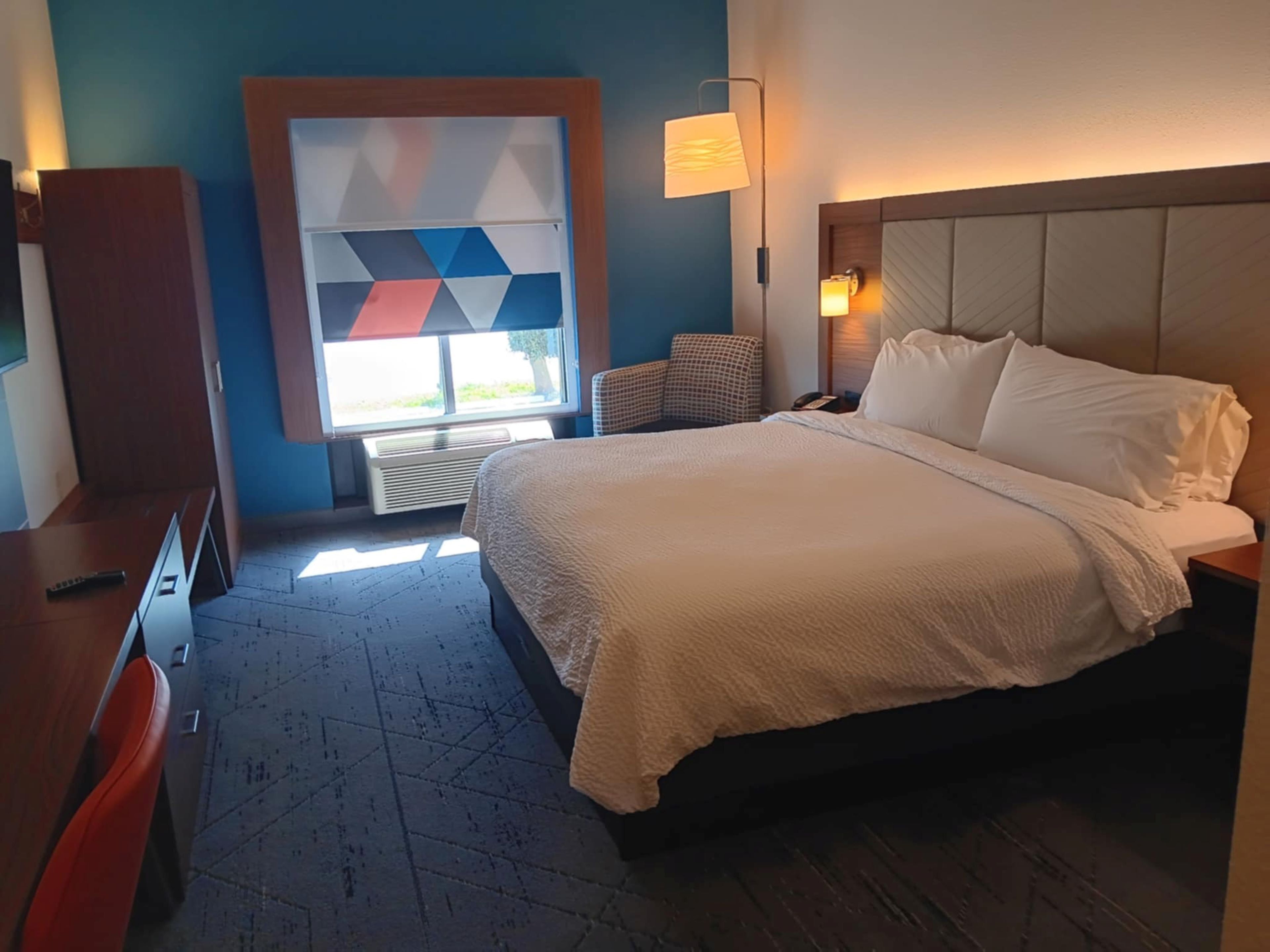 A hotel room with a bed, a desk, a chair, a wall-mounted lamp, and a window with a geometric pattern.