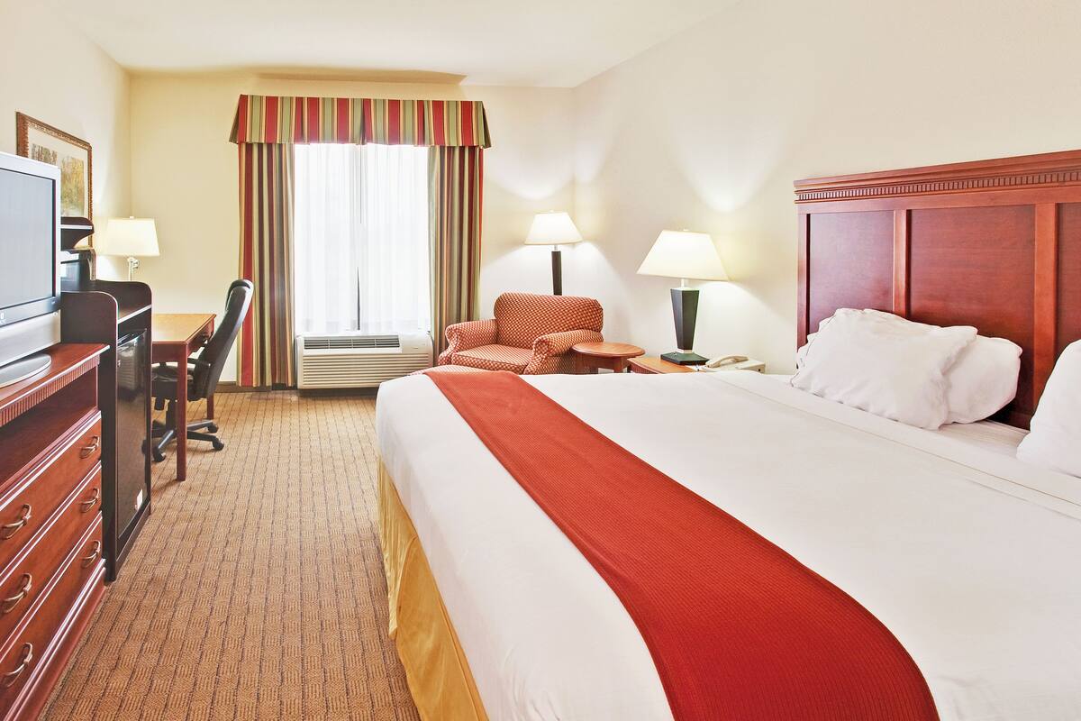 Habitación de hotel con una cama grande, un escritorio con silla, televisor, dos lámparas y una ventana con cortinas.